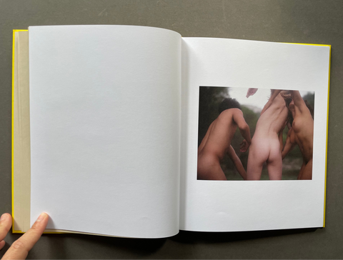 An open hardcover art book with a blank white left page and a printed photograph on the right page, held open by a hand against a dark gray background.