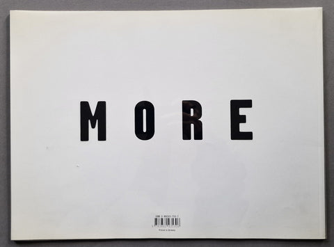 A minimalist back cover featuring the word 'MORE' in bold black sans-serif letters on a white background, photographed flat against a dark gray surface.