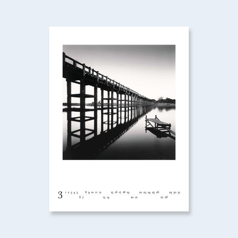 A minimalist calendar page featuring a long bridge reflected in calm water.