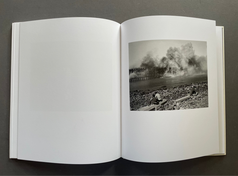 An open hardcover book laid flat on a dark gray surface, showing a black-and-white photographic print on the right page depicting a smoky industrial scene near a rocky shoreline.