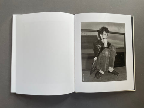 An open hardcover book lying flat on a dark gray surface, showing a large blank white left page and a black-and-white photograph on the right page of a seated person.