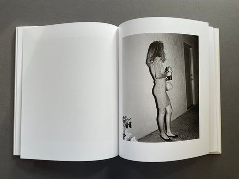 An open hardcover book laid flat on a dark gray surface, showing a black-and-white interior photograph printed on the right page. The photo depicts a woman standing in a hallway holding a bottle.