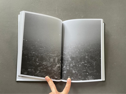 An open hardcover book displaying a black and white aerial photograph of a dense cityscape, held open by two fingers against a dark gray background.