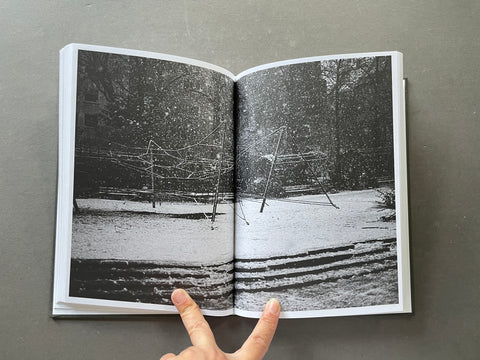 An open hardcover book showing a black and white photograph of a snowy playground scene, held open by two fingers against a dark gray background.