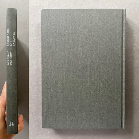 A hardcover book with a textured gray linen cover, shown from the front and spine. A hand is holding the spine on the left to show the binding and text.
