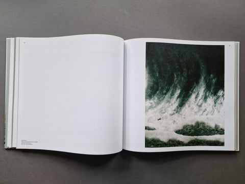An open art book, showing a large white left page and a right page with a dramatic dark green and white wave photograph.