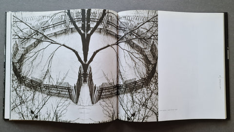 An open book laid flat on a dark surface, showing a black-and-white symmetrical photograph of a tree and fence on the left page and blank white page space on the right.
