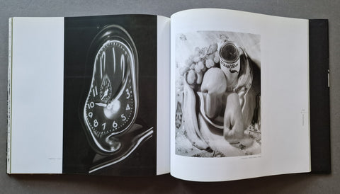 An open art book spread on a dark surface, showing two black-and-white photographic prints: a surreal melting clock on the left page and a monochrome still-life still life on the right page.