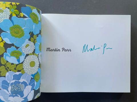 An open book or journal showing a white interior page with the printed name Martin Parr and a blue ink signature, next to a floral-patterned cover in blue, green, and gray.