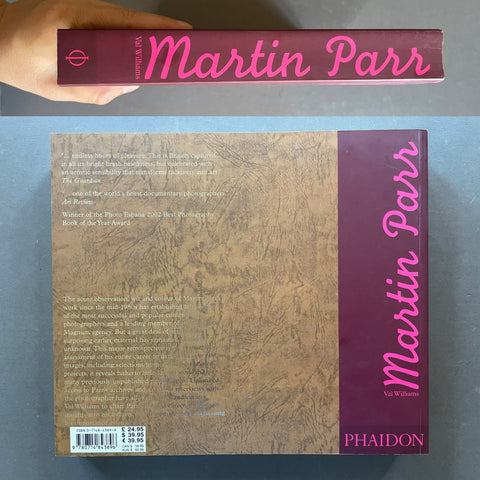 A hand holds a hardcover book titled 'Martin Parr' above the back cover of the same book on a gray surface. The cover features a textured brown background with a dark purple vertical band on the right side containing the title in pink script and the PHAIDON logo at the bottom.