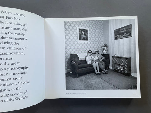 An open book or magazine spread showing a black-and-white interior photograph of a couple sitting on a sofa in a living room, with printed text visible on both sides of the page against a dark background.