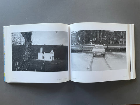 An open hardcover book laid flat on a dark gray surface, showing two black-and-white photographic prints on the pages. The left page features a small white house in a dark landscape, and the right page shows a vintage car on a wet street with buildings in the background.