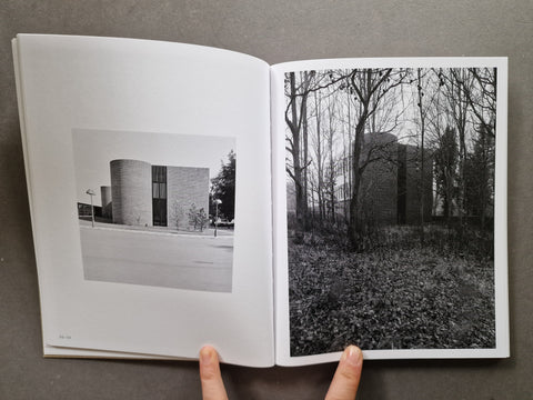 An open book or magazine spread showing two black-and-white architectural photographs on the pages, held at the bottom by two fingers against a gray background.