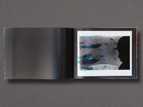 An open art book  laid flat on a dark gray surface, showing a black-and-white silhouette portrait in a square print alongside an abstract blue-toned photographic page.