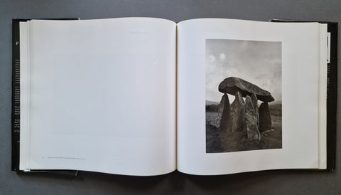 An open book showing a black-and-white photograph of a stone megalithic structure on the right page, with blank white pages on the left, photographed from above on a dark gray surface.