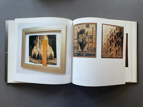 An open art book displayed flat on a dark gray surface, showing two interior spreads with framed and printed artwork. The left spread features a square abstract composition with black, white, and orange tones inside a light wood frame, while the right spread shows two sepia-toned architectural or figurative prints.