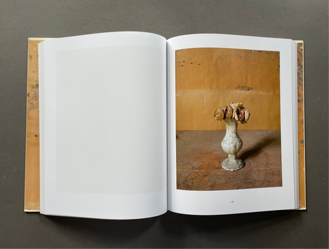 An open art book or photo journal lying flat on a dark gray surface. The left page is blank white, while the right page features a photograph of a small white vase holding dried flowers against a warm brown background.