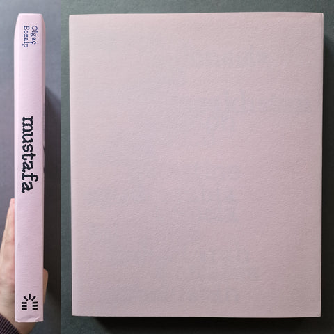 A pale pink book shown standing upright, with its spine visible on the left and a blank cover on the right. A hand is holding the spine at the lower left edge.