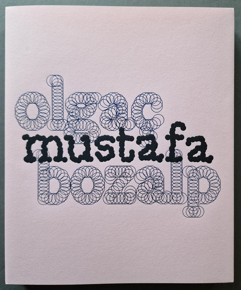 A square art book cover with the title "Mustafa" and the name "Olgac Bozalp" in stylized black and dark blue stitched-texture lettering on a pale pink paper background, photographed against a dark green surface.