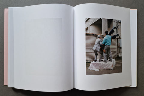 An open book, with mostly blank white pages on the left and a printed photograph on the right showing two people standing outdoors near a corrugated metal wall.