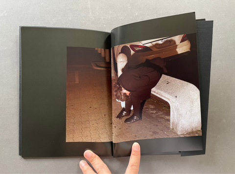 An open black photo book or magazine laid on a gray surface, showing a printed photograph on the right page of a person in dark clothing bending over near a bench at night. A hand is visible holding the book open.
