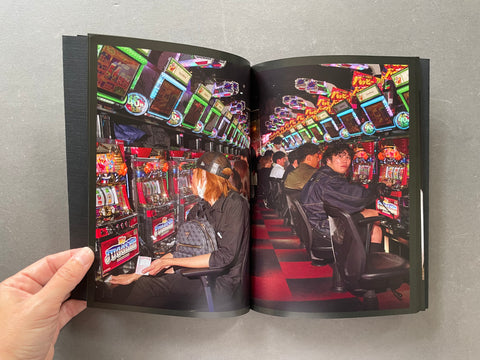 An open book or magazine spread showing a color photograph of people sitting at brightly lit slot machines in a casino-like interior, held open by a hand against a gray background.