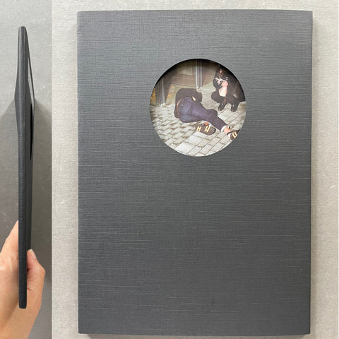 A dark gray rectangular case or book-like product with a circular cutout showing an illustration, photographed on a light gray surface. A hand holds a matching slim black cover or insert at the left edge.