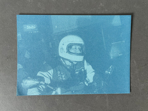 A square art print or postcard featuring a blue-toned photograph of a person in a white helmet, placed on a dark gray textured surface.