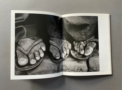 An open book or magazine spread showing a black-and-white photographic print of bare feet wearing sandals, photographed on a smooth gray surface.