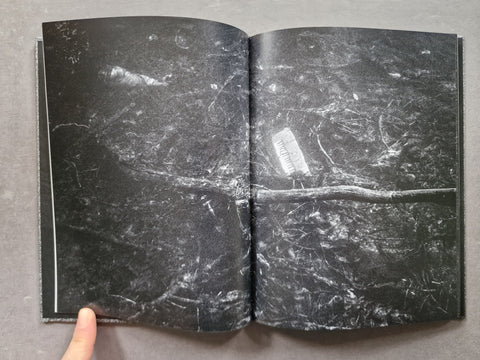 An open book showing a dark, textured black-and-white surface with a white rectangular object placed on top, photographed from above on a gray surface with a finger visible at the bottom left.