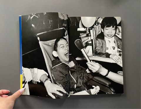 An open vintage black-and-white photo book held open by a hand, showing a candid photograph of children in wheelchairs with a microphone in a dim indoor setting, photographed against a dark gray background.