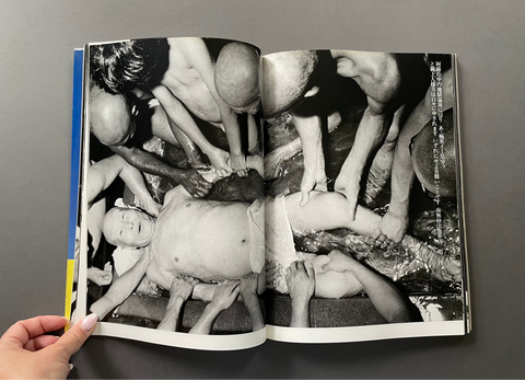 An open book showing a black-and-white photographic print of multiple people gathered around a person on the ground, photographed against a gray background with a hand holding the left page.