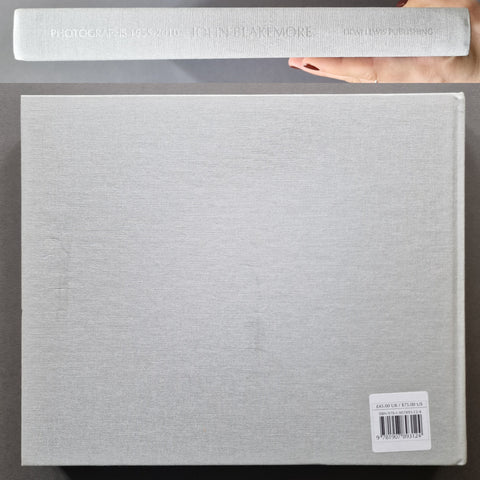 A hardcover art book or photo album shown from the back, with a hand holding the top edge of the spine. 