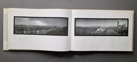 An open vintage book spread on a dark gray surface, showing two black-and-white panoramic landscape photographs printed on white pages.
