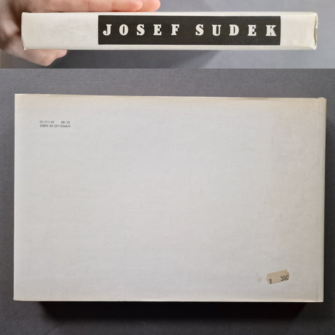 A book titled 'JOSEF SUDEK' is shown with its front cover open or laid flat below, held at the top by a hand. The cover is plain off-white with black title text and small printed text in the upper left corner.