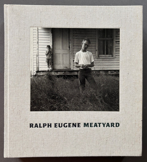 Book titled 'Ralph Eugene Meatyard' by Ralph Eugene Meatyard with a beige cover featuring a black and white photograph of 2 boys in front of a wooden house.