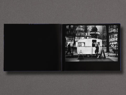 An open black-and-white photo book lies flat against a dark gray background. The left page is solid black, while the right page shows a black-and-white street photograph of a small trailer or food cart with pedestrians and storefronts in the background.