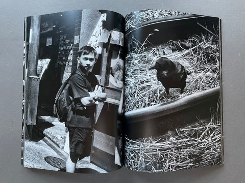 An open black-and-white art book or photo print showing two photographic scenes: a person standing in a doorway on the left page and a dark bird in straw on the right page, photographed against a plain gray background.