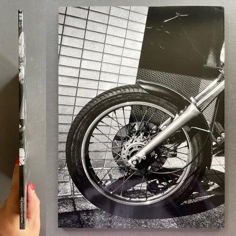 Close-up of a motorcycle wheel on a tiled floor with a hand holding a book.