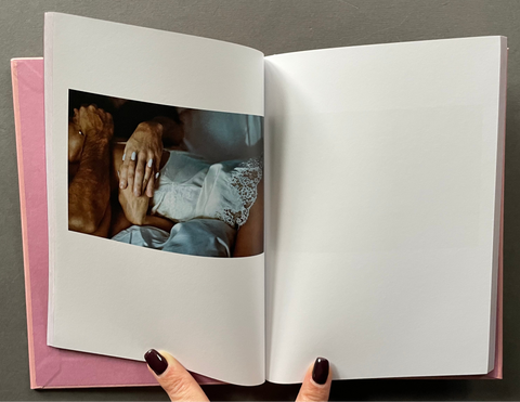 An open hardcover book or photo album lies on a dark gray surface. The left page shows a printed photograph of hands holding a baby wrapped in white cloth, while the right page is blank white.