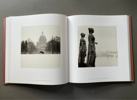 An open hardcover book laid flat on a dark gray surface, showing two printed black-and-white photographic panels on the spread. The left page contains a sepia-toned image of a domed building, and the right page contains a monochrome image of two statues by water.