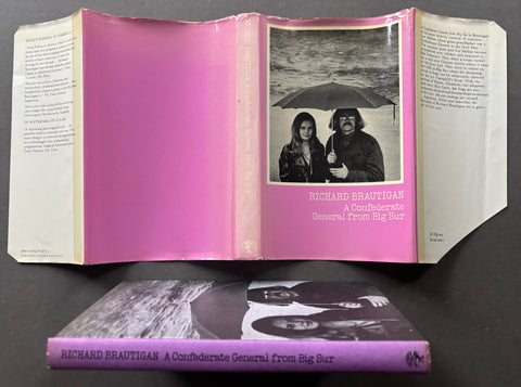 An open vintage hardcover book titled 'Richard Brautigan: A Confederate General from Big Sur' displayed above its closed cover, both resting on a dark surface. The book's cover and interior pages feature a prominent pink background with a black-and-white photograph of two people under an umbrella.