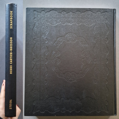 A black embossed leather-bound scrapbook book shown from the front cover and spine, held by a hand on the left. The spine reads 'SCRAPBOOK', 'HENRI CARTIER-BRESSON', and 'STEIDL' in gold lettering.