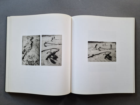 An open book showing two black-and-white photographic prints on the left page and one larger black-and-white print on the right page, photographed from above on a dark gray surface.