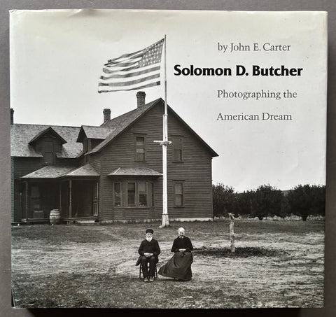 Book titled Photographing the American Dream' by Solomon D. Butcher with a black and with cover featuring the image of a man and woman sitting outside a house and a flagpole flying the stars and strips.