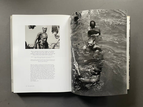 An open book spread showing a black-and-white photograph of people in the water on the right page and a smaller inset photo with text on the left page, photographed from above on a dark gray surface.