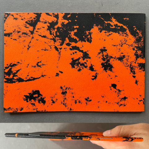 An orange and black abstract patterned notebook or journal is shown standing upright on a gray surface, with a hand holding a matching slim notebook at the left edge.