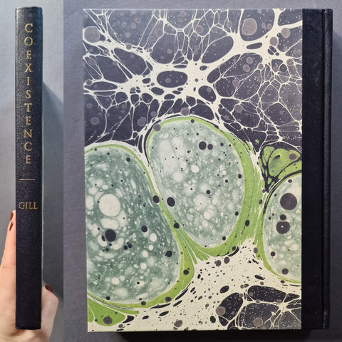 A hardcover book titled 'COEXISTENCE' by Gill, shown standing upright next to a hand holding the spine. The cover features a marbled abstract pattern in green, black, and off-white, with gold lettering on the spine.