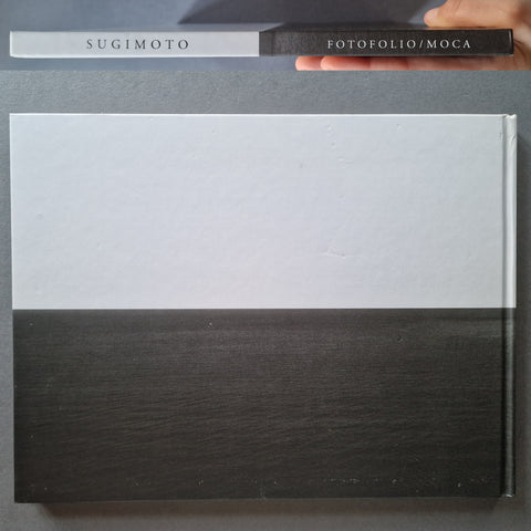 A large flat rectangular book with a white upper half and dark charcoal/black woodgrain lower half, shown on a gray surface. A smaller matching item at the top has the text 'SUGIMOTO' and 'FOTOFOLIO/MOCA', and a hand is holding it.