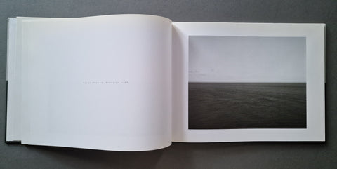 An open book showing a black and white photograph of a calm sea under a cloudy sky, placed on a dark gray surface.